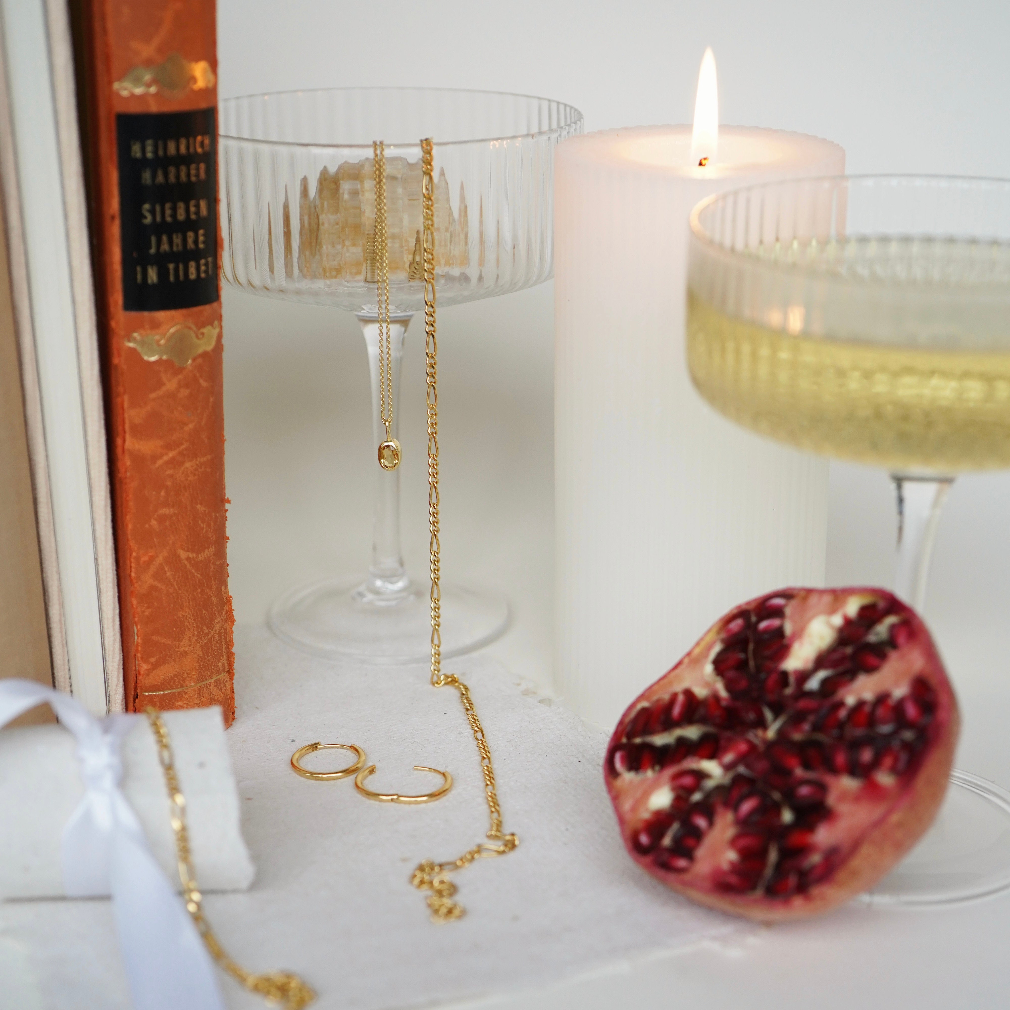 Goldfarbene Ketten an einer Glasschale neben einer brennenden Kerze und einem orangefarbenen Buch