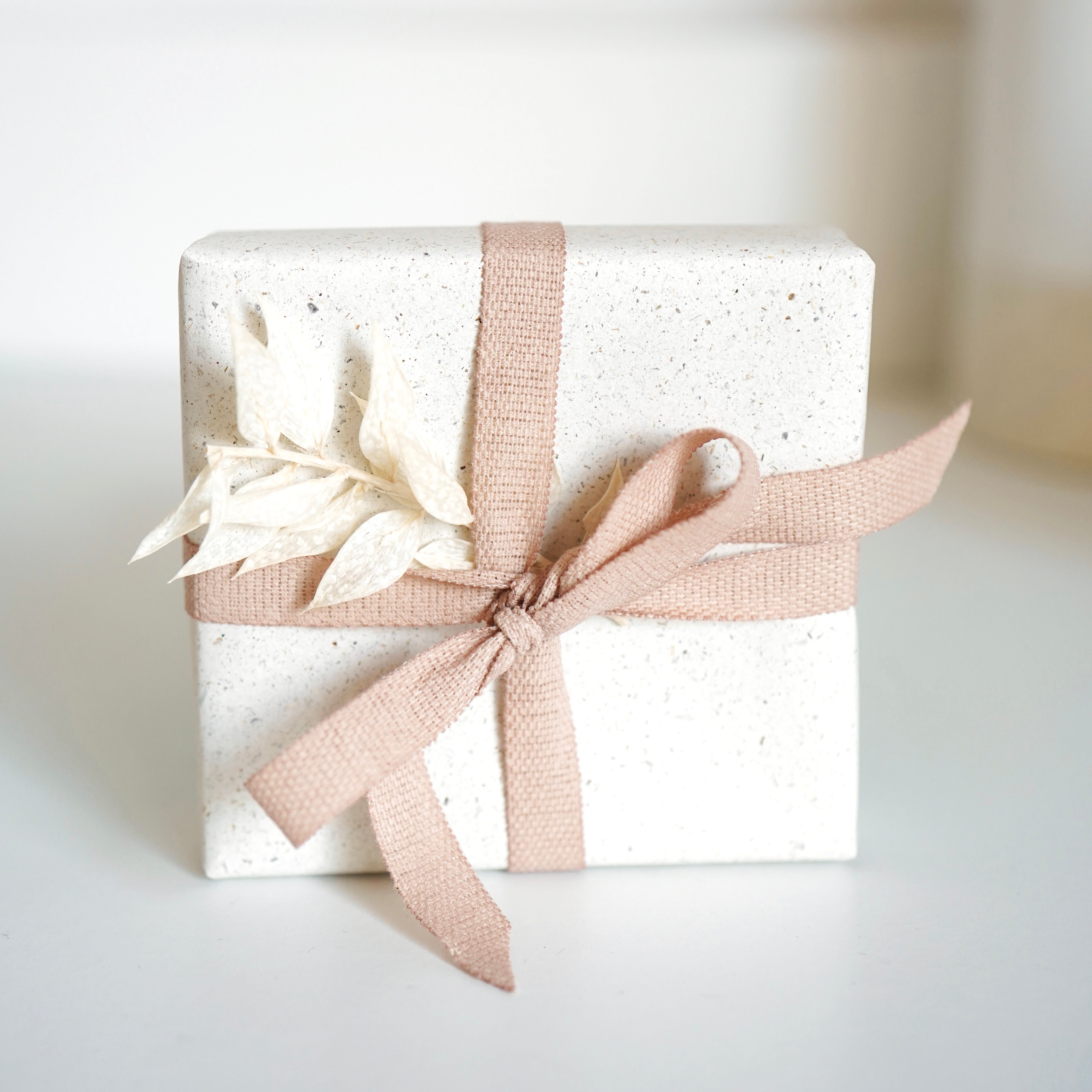 Weiße Geschenkbox mit rosafarbenem Stoffband und getrockneten Blüten